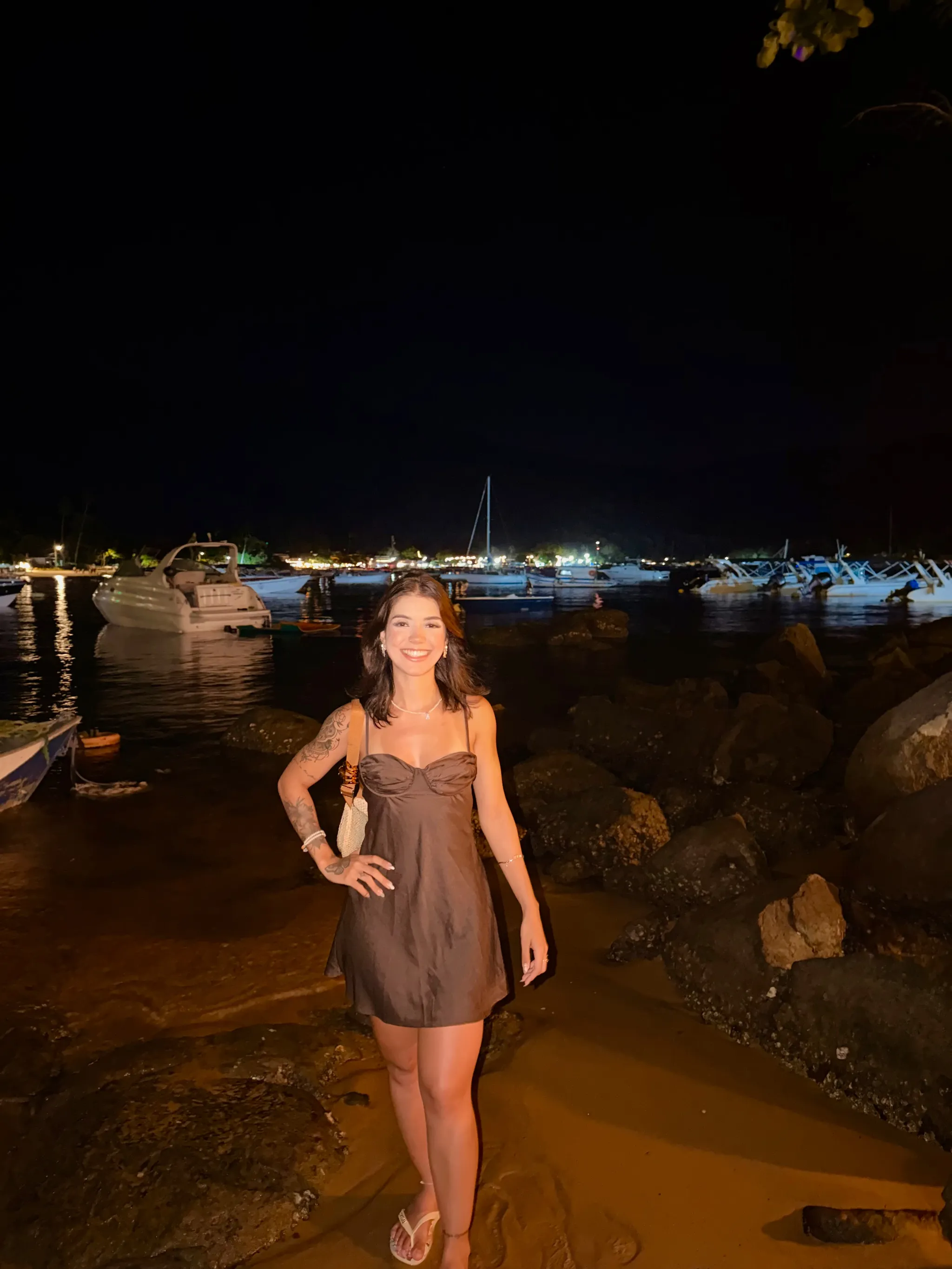 Anne Caroline standing by the water at a marina after dark, boats moored on black water and a string of warm lights strung across the far shore behind her