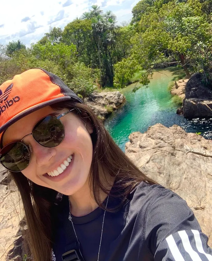 Anne Caroline in an orange cap and sunglasses, laughing beside a natural rock pool of clear green water