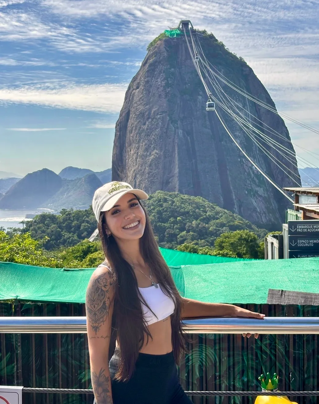 Anne Caroline smiling at the Pão de Açúcar cable car station in Rio de Janeiro, the mountain and the bondinho cables behind her